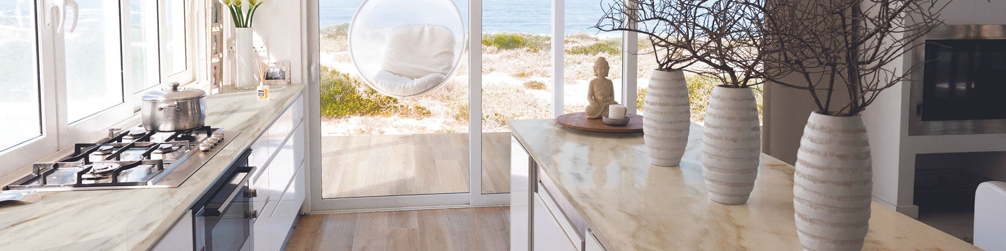 Long kitchen island with beige quartzite countertop, holding large vases with dried twigs, and view of beach & ocean.