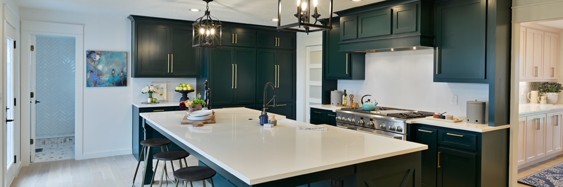 Remodeled kitchen with white quartz countertops and backsplashes, dark green cabinets with brass hardware, and black cage pendants over island.