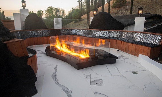 Jason Derulo’s backyard with glassed-in firepit, wooden bench seating with black glass mosaic tile, flooring of white & gray porcelain tile that looks like marble.