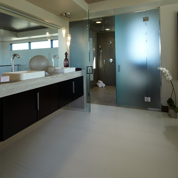 Bathroom with beige limestone waterfall countertop on floating double vanity, pendant lighting, 12x24 fabric look tile flooring, and wet room with brown floor & wall tile.