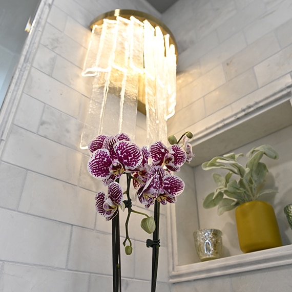 Closeup of wall sconce on white & gray marble bathroom backsplash, violet orchid, and succulent & candle in wall niche.