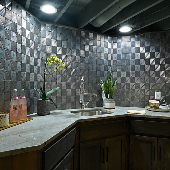 Basement bar with silver textured geometric wall tile that looks like metal tile and beige quartzite countertop over natural wood cabinets.