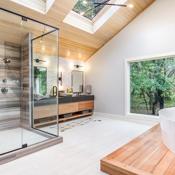 Renovated bathroom with steam shower, brown tile that looks like heavily striated marble, marble mosaic backsplash, brown quartz vanity countertop, and vaulted wood ceiling with skylights.