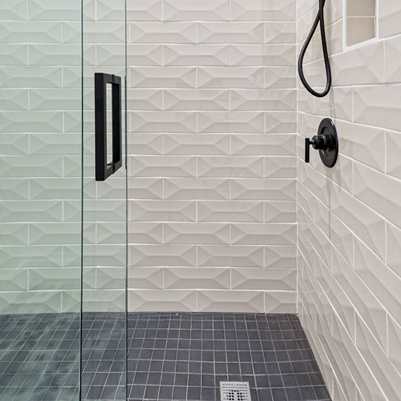 Renovated shower with white textured wall tile, black fixtures, black mosaic shower floor tile, and frameless shower glass with black handle.
