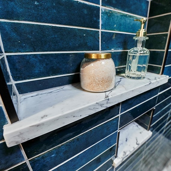 Closeup of shower niches with blue glazed porcelain wall tile, marble shelf holding bath salt & glass liquid soap dispenser.