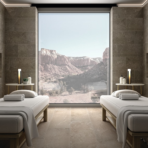 Massage room with tan floor and wall tile that look like stone, wall niches holding candles, and large picture window with view of desert plateau between two massage tables.