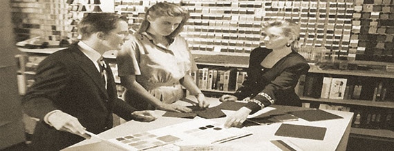 Black and white photo of a man and two women looking at tile samples on a table.
