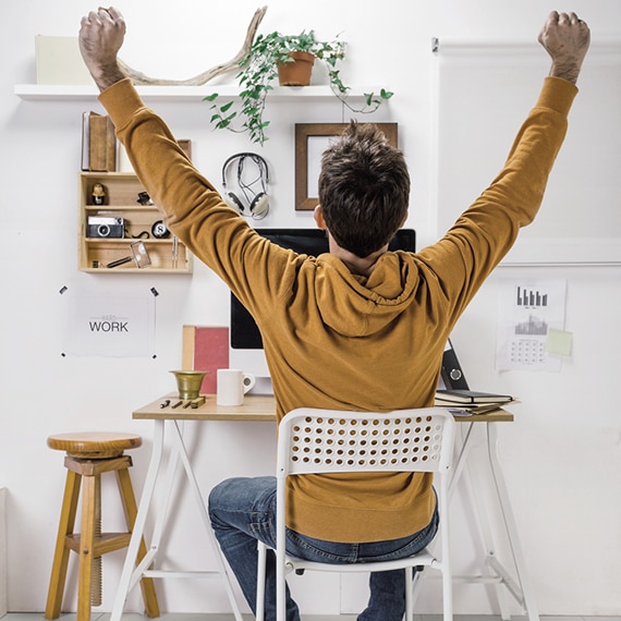 Back view of a teenaged male, sitting at a computer desk & chair, holding up his arms in celebration.