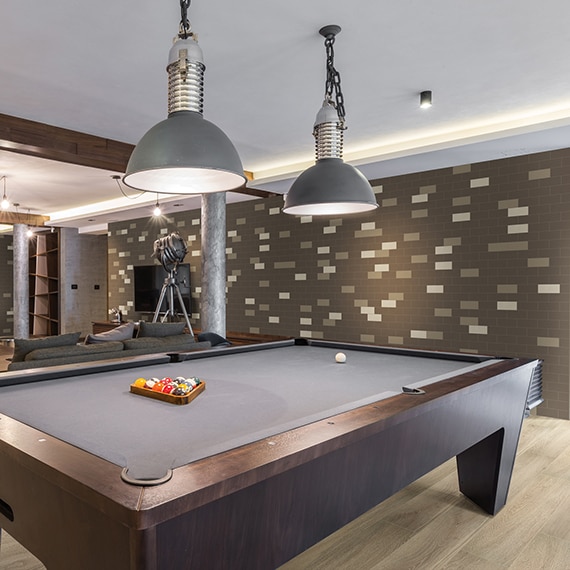 Game room with gray felt billiard table, floor tile that looks like wood, white, tan & brown subway tile, wood beam ceiling, and brown over-stuffed couches.