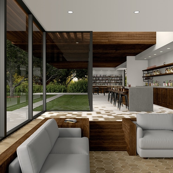 Restaurant lobby with tan stone look hexagon floor tile, gray couches, gray quartz bar top, black barstools, floor-to-ceiling windows revealing lawn and walkway of white pavers.