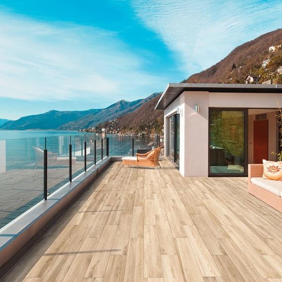Solarium with wood look tile deck & glass railing overlooking a coastline with blue skies and mountains in the background.