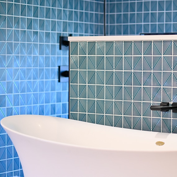 Closeup of blue geometric shower tile and wainscot tile behind a free-standing white bathtub with black faucet.