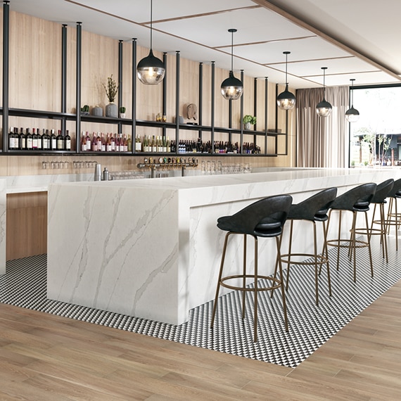 Hotel bar with white & gray marble look quartz countertops, black & white checkered tile mat set in wood look tile flooring, and racks holding wine bottles in front of wood panel wall.