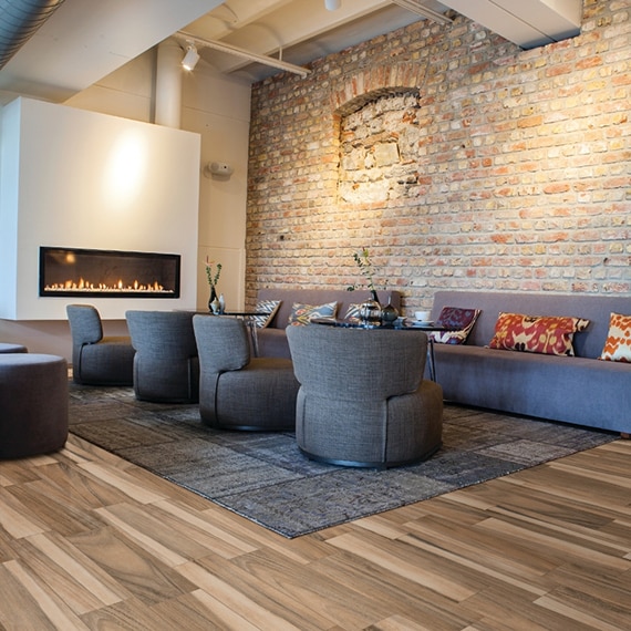 Lobby with wood look tile floor, blue area rug, chairs, and sofas, wall mounted electric fireplace, and brick wall.