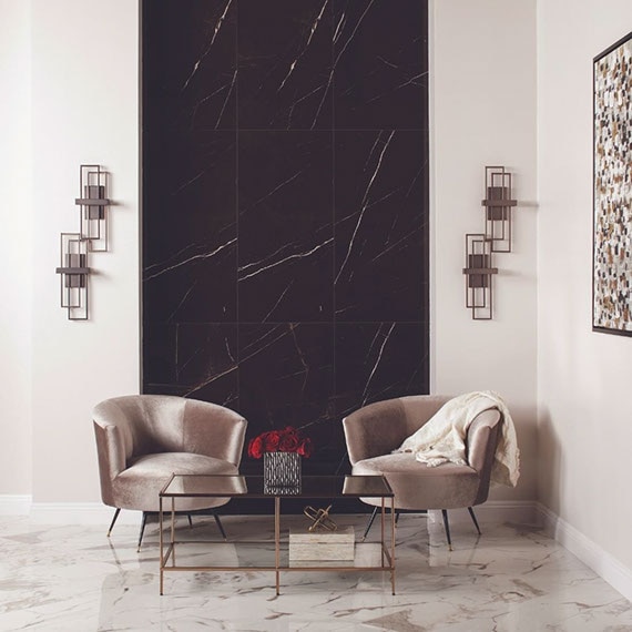 Lounge area with white & gray floor tile and black wall tile that look like marble, beige velvet chairs behind brass and glass coffee table.