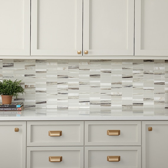 Off-white kitchen cabinets, off-white & beige marble mosaic backsplash, off-white & gray veining quartz countertop, plant in terracotta pot on top of two books.