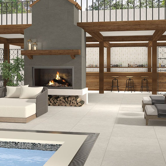 Hotel pool with gray stone-look deck tile, outdoor fireplace with gray tile surround and natural wood mantle, off-white chaise lounge chairs, and outdoor bar.