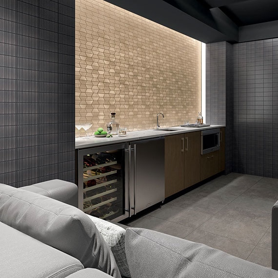 Home theater with gray concrete floor tile, charcoal metallic look wall tile, wet bar with brass trapezoid mosaic backsplash, ivory quartz countertop, and stainless steel wine refrigerator.