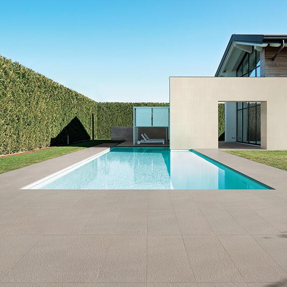 Residential pool with brown textured stone-look tile, surrounded by tall hedge, and pair of white lounge chairs with rollers.