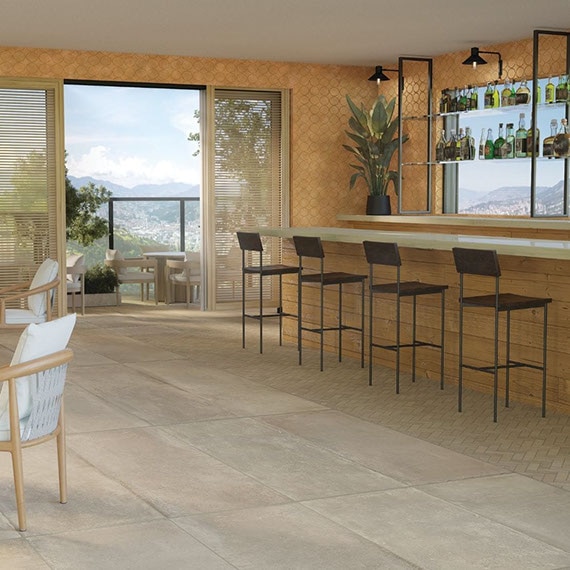 Restaurant with a mountain view, beige large-format floor tile and matching mosaic that look like concrete, woodgrain bar with bar stools, table & chair with white cushions.