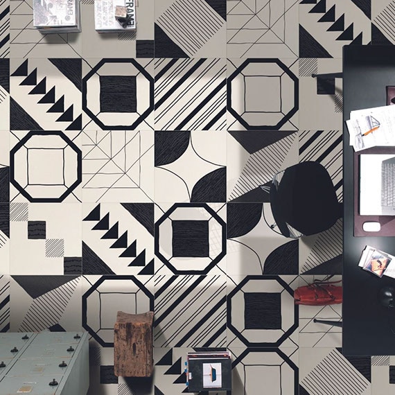 Bird’s eye view of office with white floor tile with large black designs that look hand-drawn, black metal desk & chair, gray metal lockers, and wooden table.