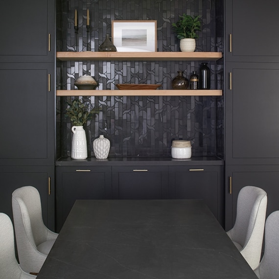 Black built-in cabinets with black marble mosaic backsplash and natural wood floating shelves in front of black quartz table with gray chairs.